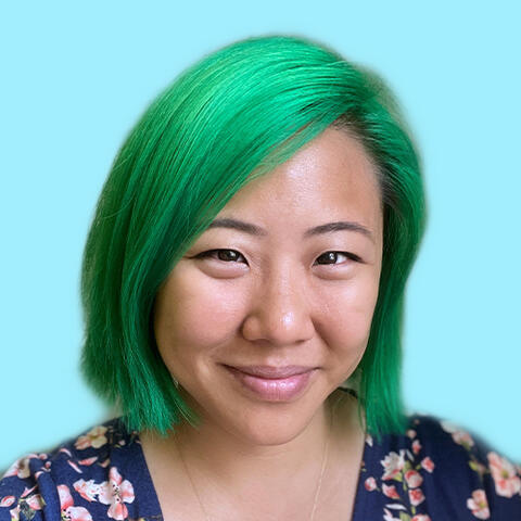 East Asian woman with shoulder length green hair looks at the camera with a closed mouth smile