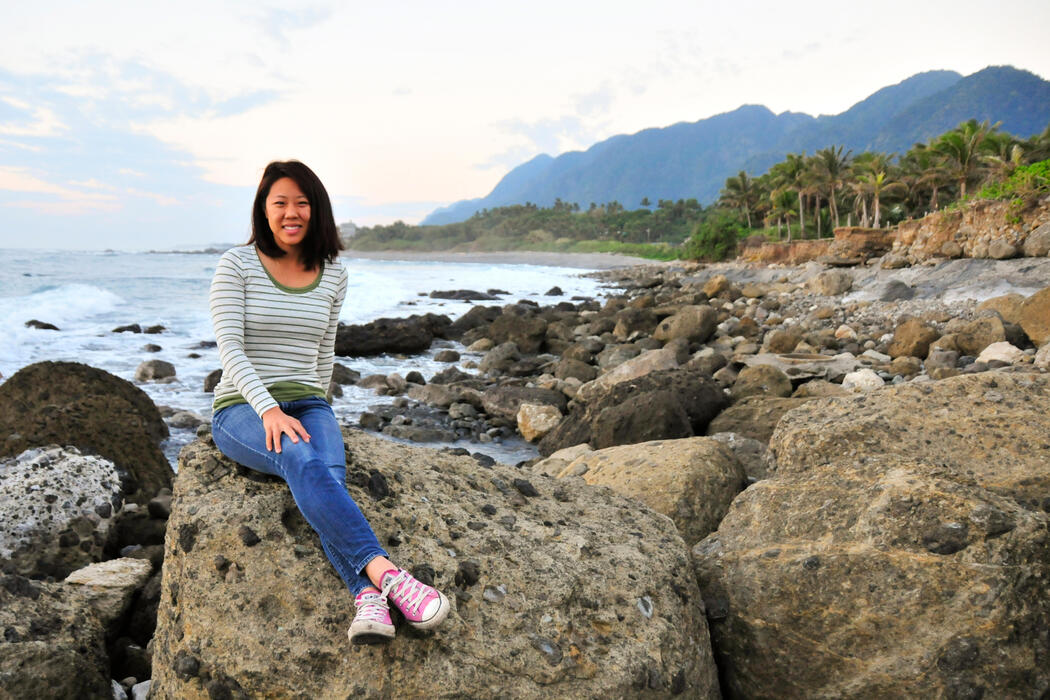 East Asian woman wearing a long sleeve shirt, jeans, and pink converse sits on a rocky beach in Taiwan with palm trees and mountains in the background